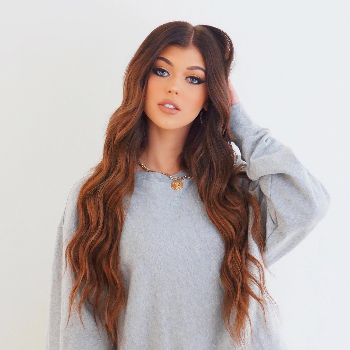 a young woman with long, wavy brown hair and blue eyes is posing indoors in a studio. she is wearing a light gray sweater and a gold necklace. she is looking at the camera with a slight smile and has her hand in her hair. the background is a plain white wall.