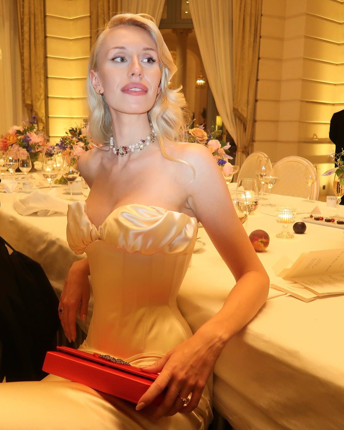 a beautiful blonde woman in a formal ivory dress and a red clutch bag, sitting at a table in a luxurious restaurant setting. she is smiling and looking toward the viewer. she has a necklace on with a red stone in the center.  the background features a dining table with more place settings,  a white tablecloth and crystal glasses, and a floral arrangement. the overall atmosphere is elegant and upscale.