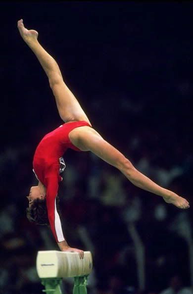 a female gymnast in a red leotard performs a graceful backbend on a balance beam. she is in midair, her body arched perfectly. the beam is made of wood and is resting on two metal stands. the background is blurred, suggesting that this is a competition or a training session.
