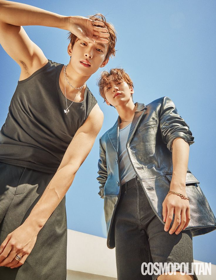 two men are posing outside for a photoshoot, smiling and laughing together. they are wearing trendy summer fashions. they are wearing black shorts, tank top, leather jacket and a white shirt. the sky is bright blue and the ground is white, giving off a summery vibe.