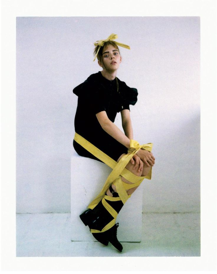 a young woman with a yellow ribbon in her hair is sitting on a white cube. she is wearing a black dress and black shoes, and her legs are wrapped in yellow ribbons. she is looking at the camera with a serious expression.