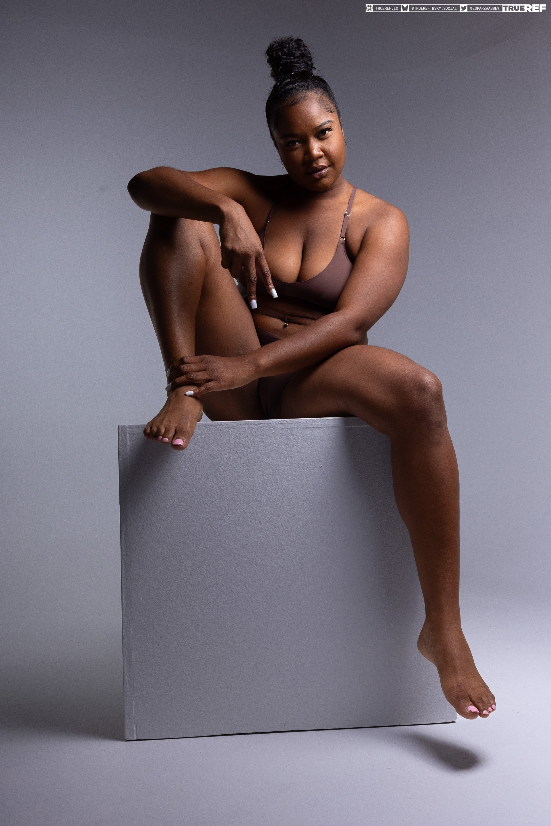 a confident and attractive darkskinned woman in brown lingerie sits atop a grey box in a studio setting. she has her hair in a bun and is posing for a medium shot against a grey background. her bare feet dangle off the box.