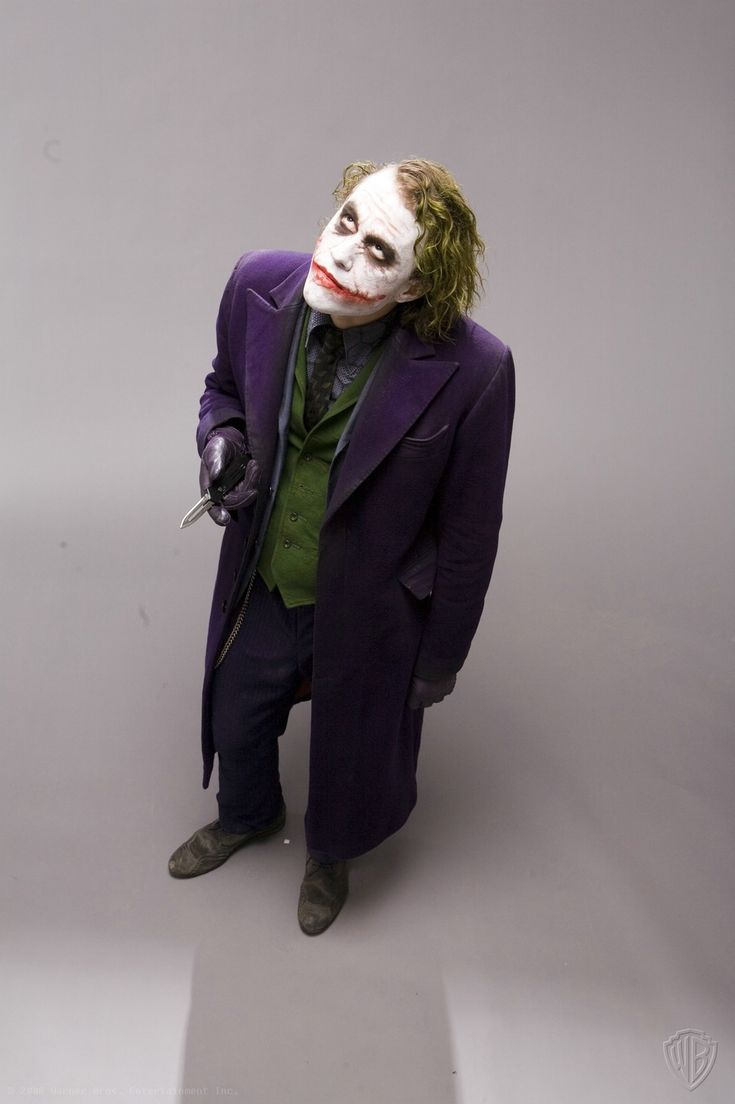 this is a photo of heath ledger as the joker from the movie the dark knight. he is wearing his iconic purple suit, green vest, and white clown makeup. he is holding a knife in his right hand and has a slight smile on his face. the background is a simple white studio setting.