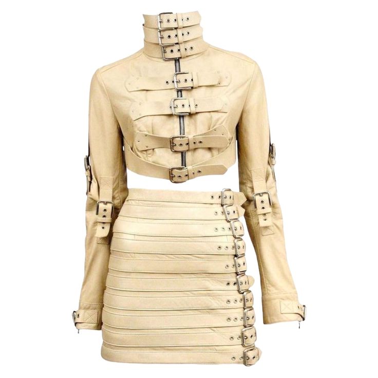 a beige leather jacket and skirt set with multiple buckles and straps. the jacket is cropped and has a high neckline. the skirt is mini and has a fitted silhouette. the clothing is designed for a woman.