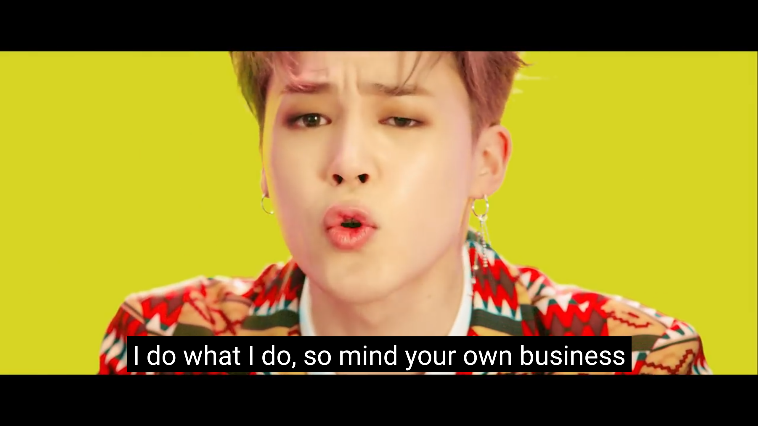 a closeup shot of bts singer jimin from the music video "boy with luv," with a yellow background and subtitles that read "i do what i do, so mind your own business." jimin has pink hair, is wearing a patterned shirt, and is making a surprised expression. he is also wearing earrings.