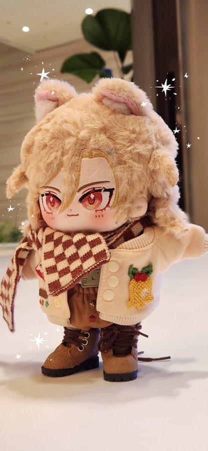 a closeup image of a plush toy dressed in a winter outfit. it is wearing a brown and white sweater, brown boots, and a brown and white scarf. it has large eyes and a sweet expression. the doll is sitting on a white surface.  the background is out of focus and features a green plant and a wall. the toy has the appearance of a character from a japanese anime or manga series.