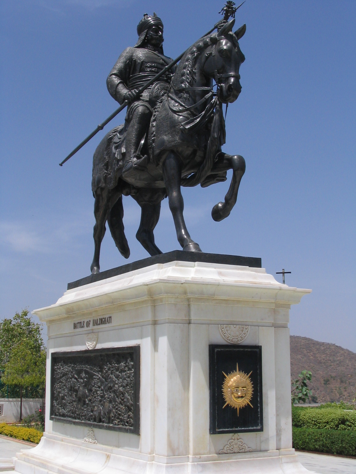 a bronze statue of a warrior on a horse stands atop a white marble plinth. the warrior holds a spear and wears traditional clothing and headwear. the plinth features decorative carvings, including a sun emblem. the background includes a clear blue sky, green trees, and a brown hill. this appears to be a monument to a historical figure.