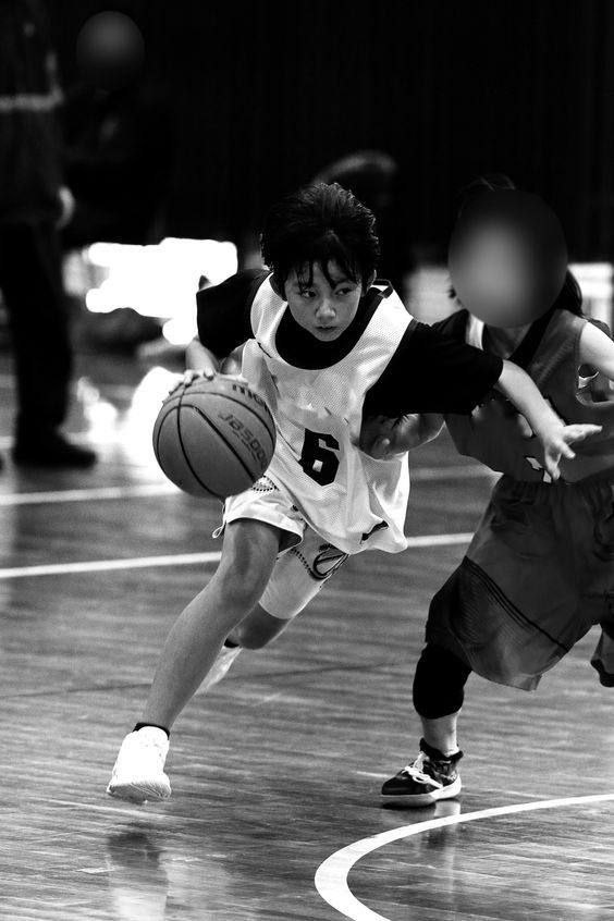 a black and white image of two female basketball players during a game. the player on the left is dribbling the ball towards the basket, while the player on the right is defending. the image is taken from an eyelevel perspective, and the lighting is bright and artificial.