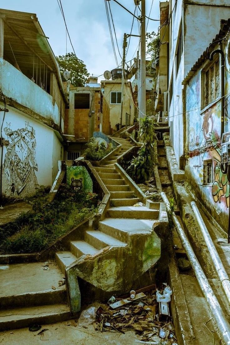 the image captures a narrow street scene, featuring a long set of concrete stairs climbing upwards between weathered buildings in a favela or slum environment. the architecture shows signs of urban decay, with graffiti and overgrown vegetation adding to the dilapidated aesthetic. the street is littered with trash, underscoring poverty. utility poles and overhead wires add to the urban clutter. the overall scene depicts the harsh realities of life in a marginalized urban area, likely in brazil.