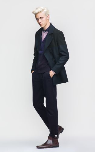 a full shot of a male model in a stylish outfit, standing in a studio against a light gray background. he has blonde hair and is wearing a doublebreasted coat, a purple shirt and vest, dark trousers, and brown chelsea boots. his hands are in his pockets, and he is posing for the camera.