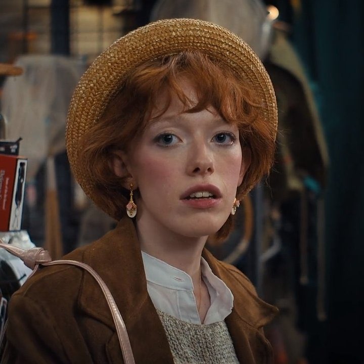 closeup portrait of a young woman with bright red hair, wearing a straw hat and a brown jacket. she has freckles and is wearing delicate earrings. she appears to be in an antique shop, with blurred background items visible.  the overall style is vintage or retro. she looks directly at the camera.