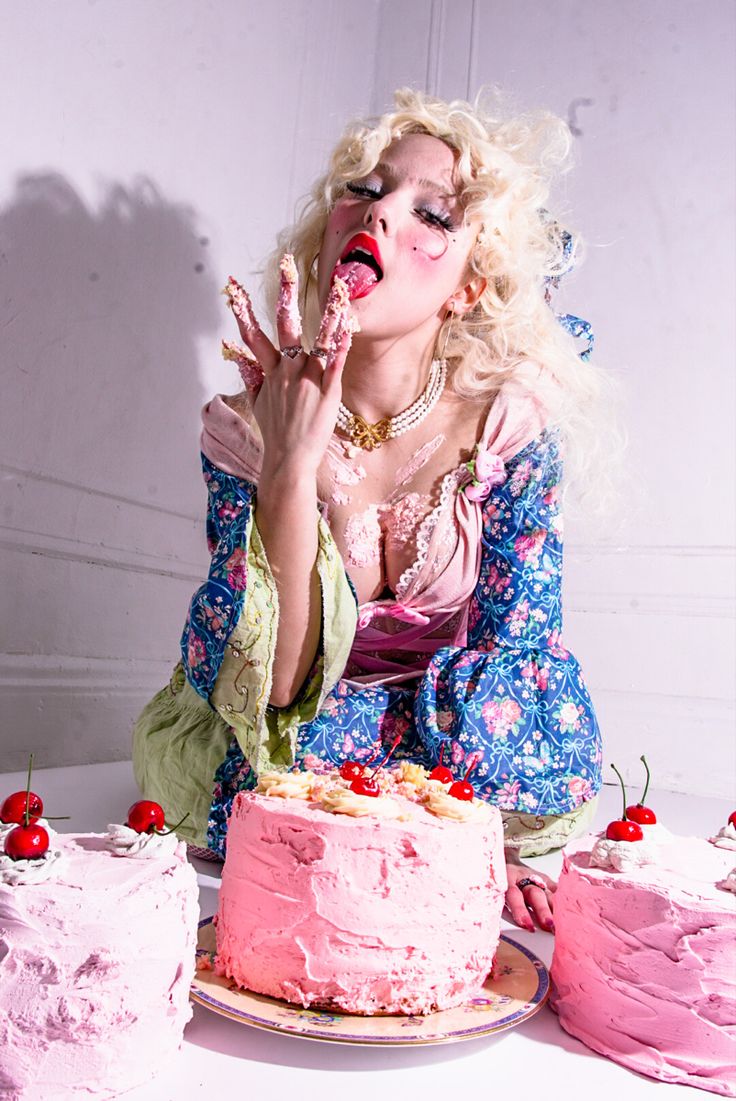 a blonde woman in a vintage floral dress is playfully eating a pink cake.  she's surrounded by several other cakes, also pink and decorated with cherries. the scene is brightly lit and styled for a fashion or art photograph, with a focus on decadent sweetness and slightly messy indulgence.  her makeup is dramatic, enhancing the overall whimsical and luxurious feel of the image.