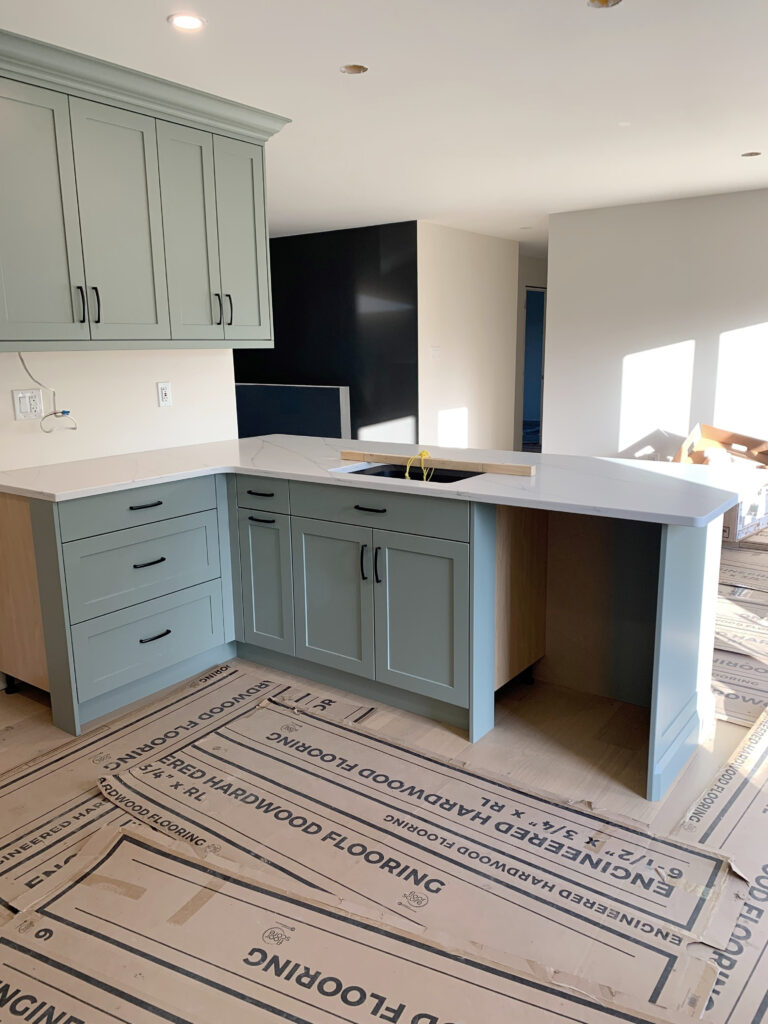 an interior shot of a kitchen under renovation. the kitchen features light blue cabinets with black hardware, a white countertop with a sink and faucet. engineered hardwood flooring, still partially covered with protective cardboard, is visible. the background reveals a dark wall feature and an adjacent lightcolored wall with sunlit areas, suggesting an open or connected space.
