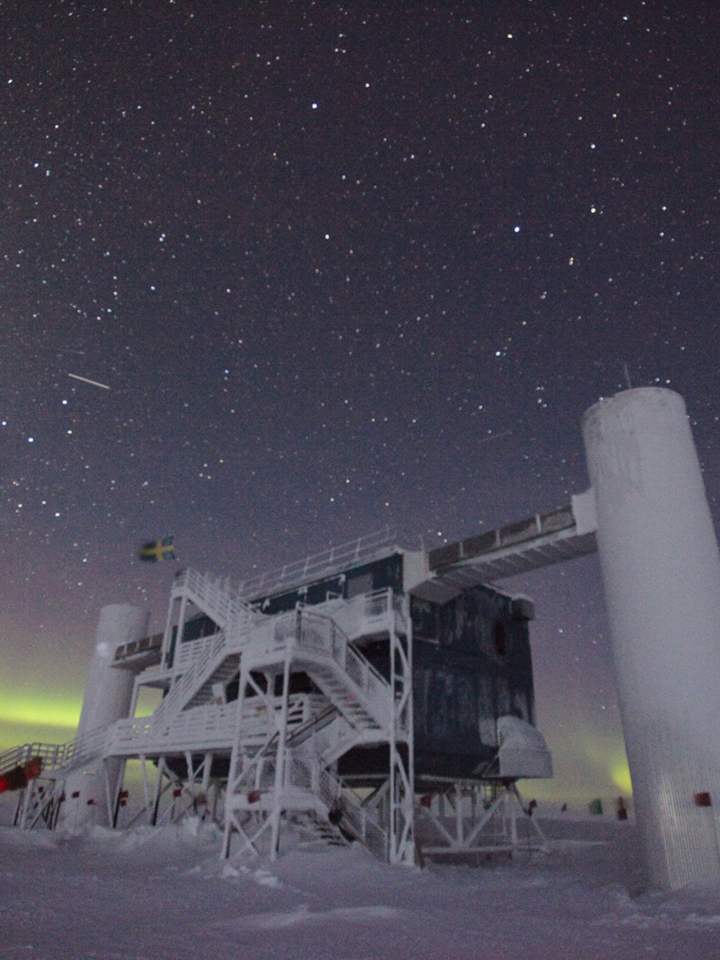 a research station in antarctica under a night sky filled with stars and the aurora borealis. the station is made of metal and is elevated on stilts. a swedish flag is flying from the top of the station. the aurora borealis is a greenish glow in the sky. the snow and ice are visible on the ground.