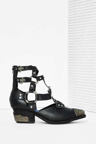 a pair of black leather ankle boots are displayed against a white background. the boots are westernstyle with a caged design, featuring multiple cutout sections and buckle straps.  they have a slight platform heel and studded details.  the boots are positioned on their side, showing the profile and details of the design.  the background is a plain white surface with a textured white wall visible in the upper right corner.