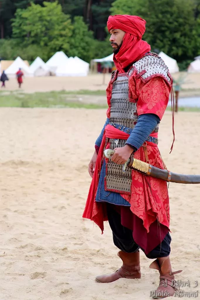 a man dressed in historical armor with a sword stands on a beach. he is wearing a red turban, metal armor over a blue shirt and red robe, black pants, and brown boots. he is holding a sword at his side. in the background there are tents and trees.