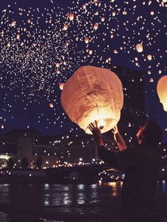 a person, possibly a woman, stands silhouetted against the cityscape, holding a glowing sky lantern in her hand. the lantern is lit up, casting a soft light, and is floating upwards into the night sky. the sky is filled with countless stars and other lanterns, creating a magical and dreamlike atmosphere. the city lights in the background are blurred and twinkling. the image evokes a sense of celebration, hope, and the pursuit of dreams.