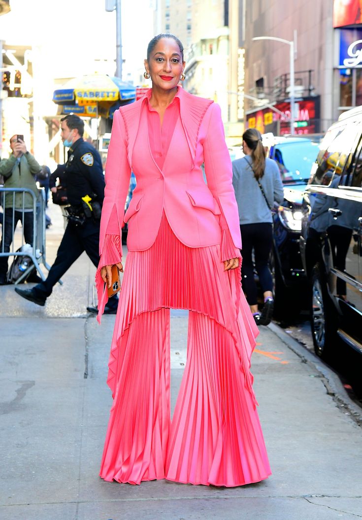 tracee ellis ross is photographed in a vibrant pink suit on an urban street. the suit features a tailored blazer with unique pleated detailing on the shoulders and fringed sleeves. the pants are widelegged and also pleated, creating a layered, cascading effect. the setting appears to be a city street during the day, with people and vehicles in the background. ross strikes a pose, looking directly at the camera.