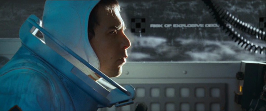 a closeup shot of a man in a futuristic blue spacesuit inside a spacecraft cockpit. he has a serious expression as he looks to the right of the frame. the cockpit is dimly lit with artificial light and features various buttons and controls. through the window behind him, a blurry, gray, lunarlike surface is visible.