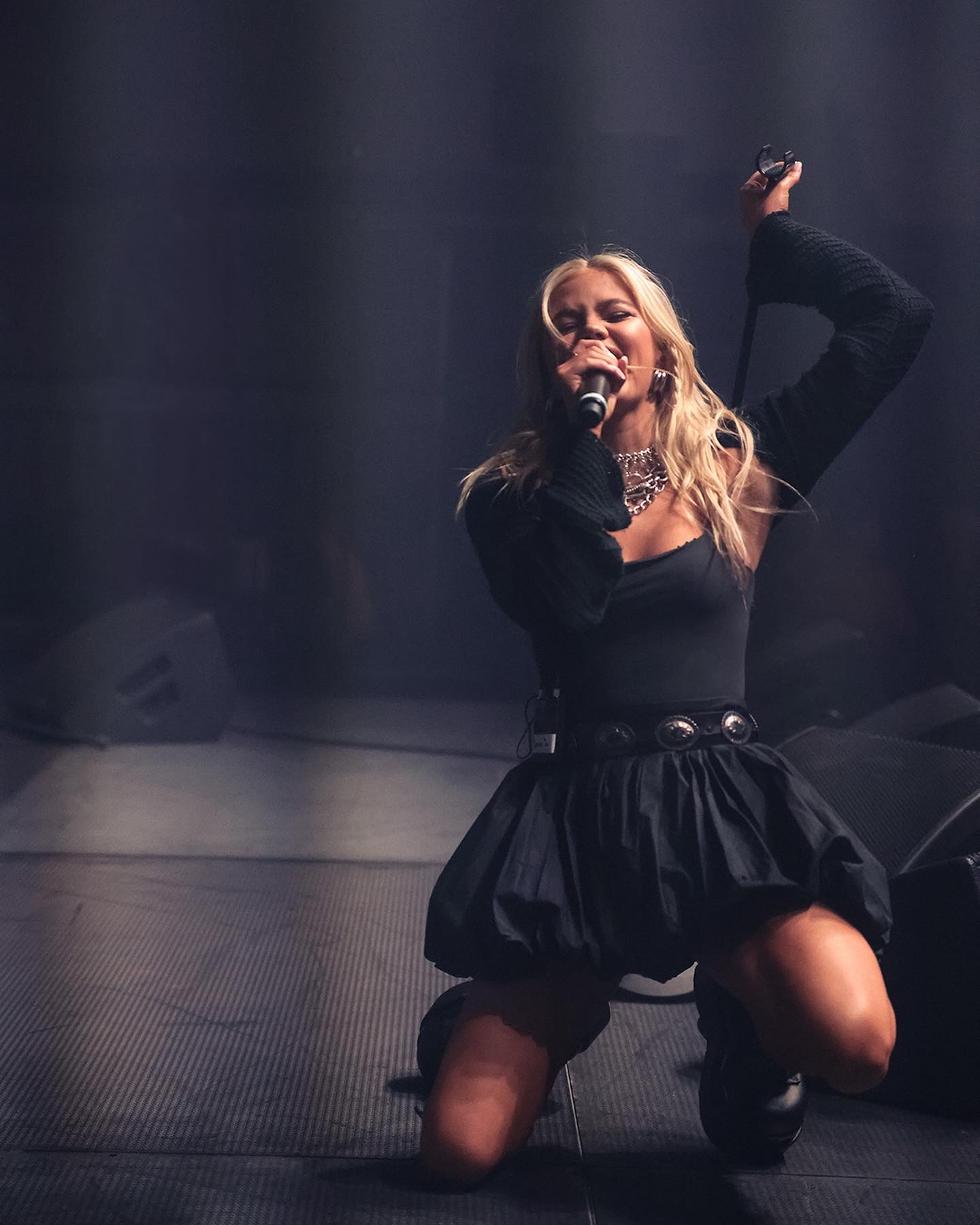 a woman is performing on stage. she is kneeling and holding a microphone while singing. she is wearing a black dress with a belt and has blonde hair. there is a dark background behind her. she is wearing jewelry. the image captures a moment from a concert or live performance.
