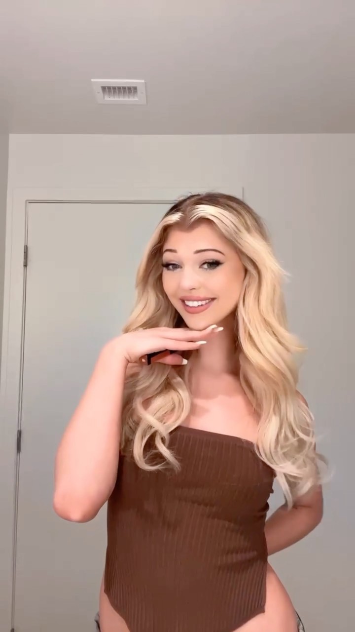 a woman with blonde hair and a brown top is smiling with her hand on her chin. she is standing indoors in a bathroom, with a door visible in the background.
