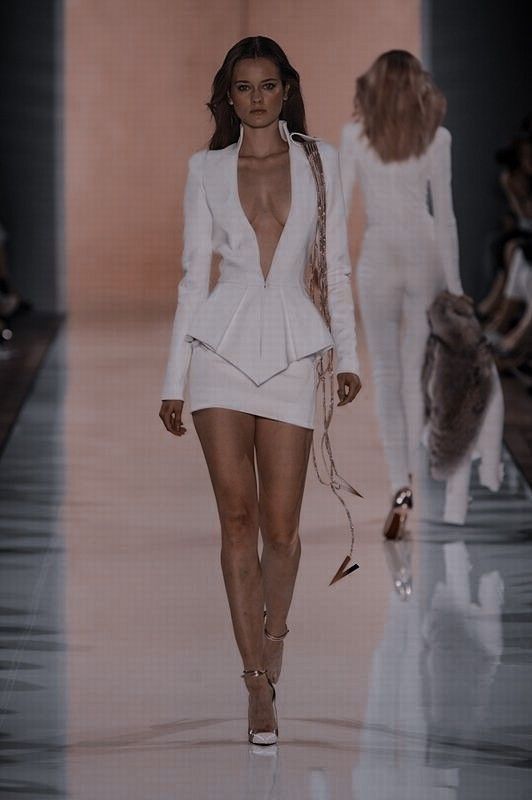 a woman in a white dress walking on a runway. the dress is short and formfitting, with a plunging neckline. she is wearing heels and has long hair. there is another model behind her in a white jumpsuit. the background is blurred.