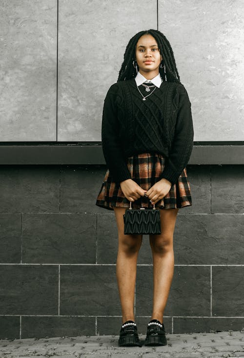 a young woman with long black dreadlocks stands confidently against a gray stone wall. she is wearing a black cable knit sweater, a brown and black plaid pleated skirt, black shoes with pearl details, and a small black handbag.  she has on a white collared shirt underneath the sweater and a simple necklace. the overall style is a mix of preppy and edgy. the image is welllit and shows her from the knees up.