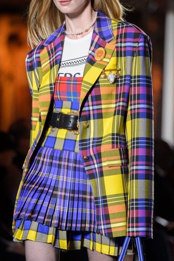 a fashion model walks the runway in a yellow and blue plaid skirt and jacket. she is wearing a white tshirt underneath the jacket. the model is also wearing a black belt and has a pearl necklace.
