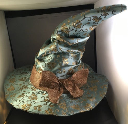 a teal and gold witch's hat with a large brim and a pointed crown, adorned with a brown bow at its base. it's crafted from a fabric with a subtle floral pattern and a slight sheen, possibly brocade. the hat rests on a surface within a box, suggesting it might be new or stored for safekeeping. the background is nondescript, allowing the hat to be the focal point.