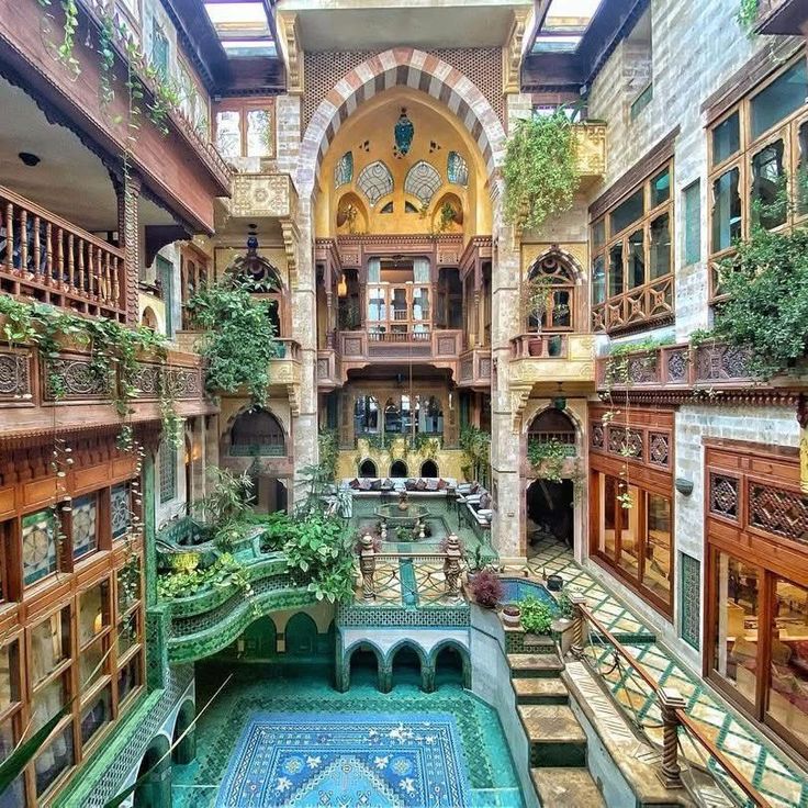 the image shows a multilevel courtyard with intricate architecture, featuring wooden balconies, arched windows, and a central fountain. lush greenery adorns the spaces, creating a vibrant atmosphere. the courtyard is richly decorated with colorful tiles and a patterned carpet, complemented by comfortable outdoor seating arrangements. a skylight above allows natural light to flood the area.