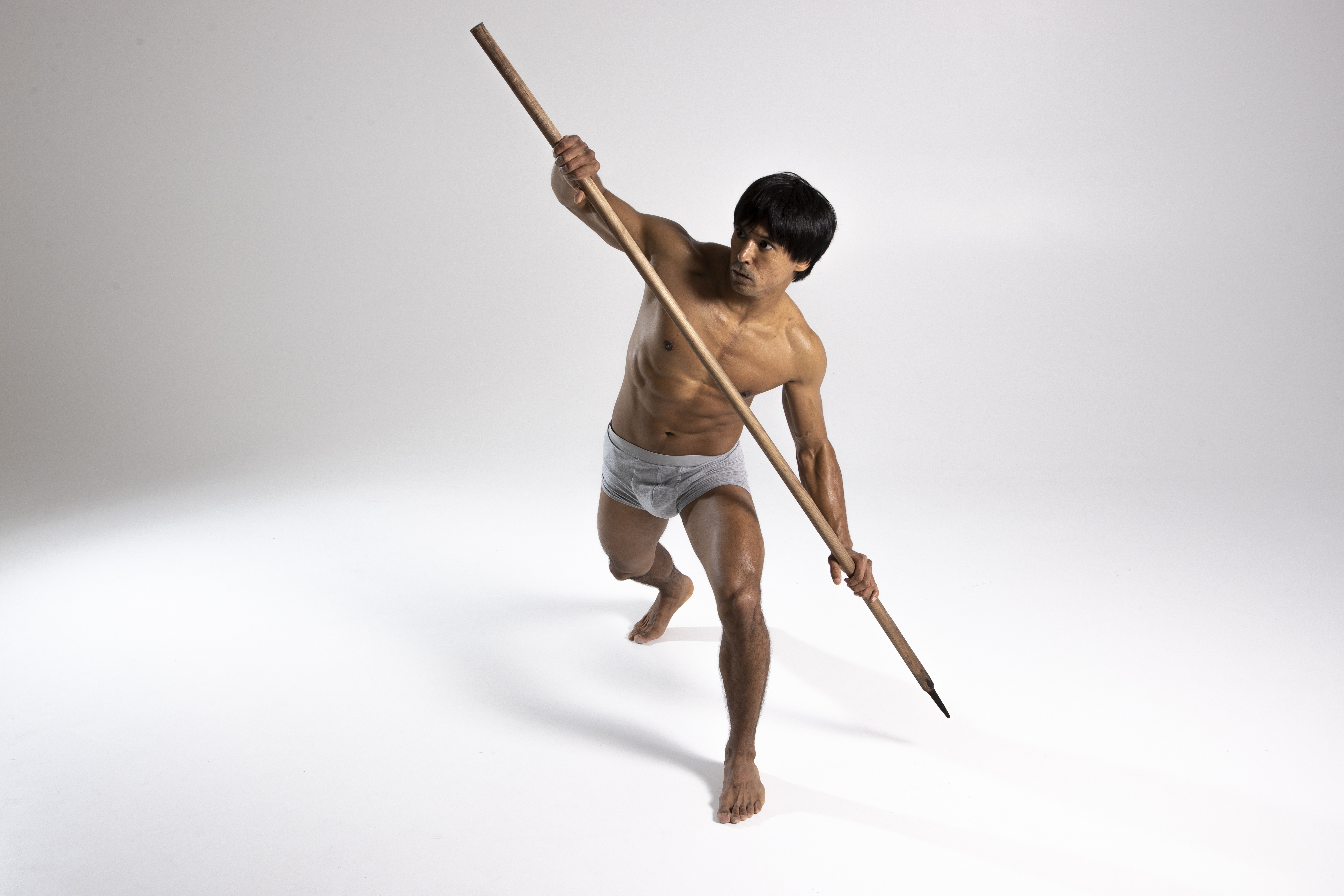 a shirtless, muscular asian man in underwear strikes a warrior pose with a spear in a studio setting. he is barefoot and looking away, exhibiting athleticism and aggression in a fighting stance. the image conveys action, defense, combat, and selfdefense, representing an active lifestyle and martial arts.