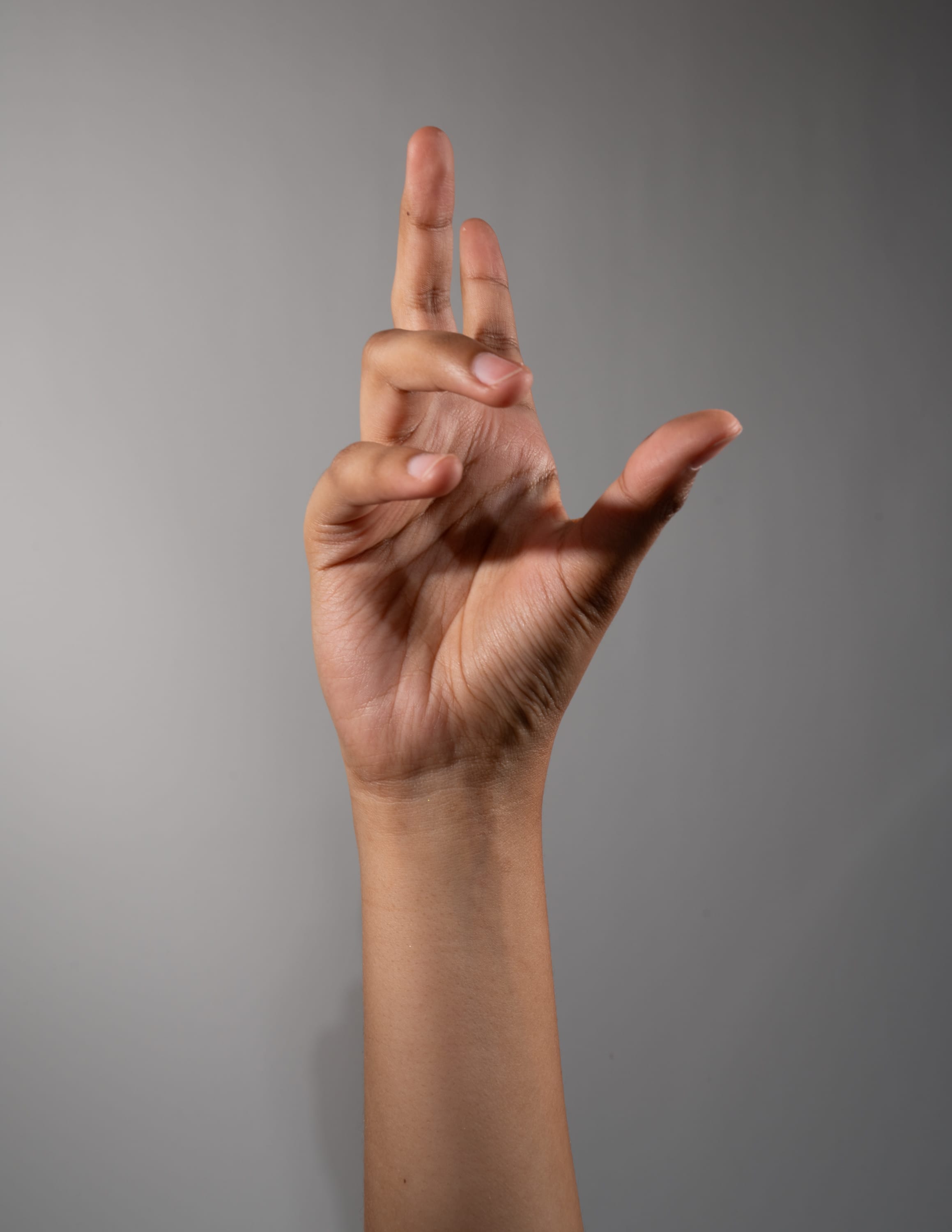 a single hand is raised against a neutral gray background. the hand, with a medium skin tone, is making a distinct gesture with the index, middle, and ring fingers extended upward, while the thumb and pinky are tucked into the palm. the pose resembles a common gesture for the number "3". the soft, natural lighting evenly illuminates the hand, highlighting its details and shape.