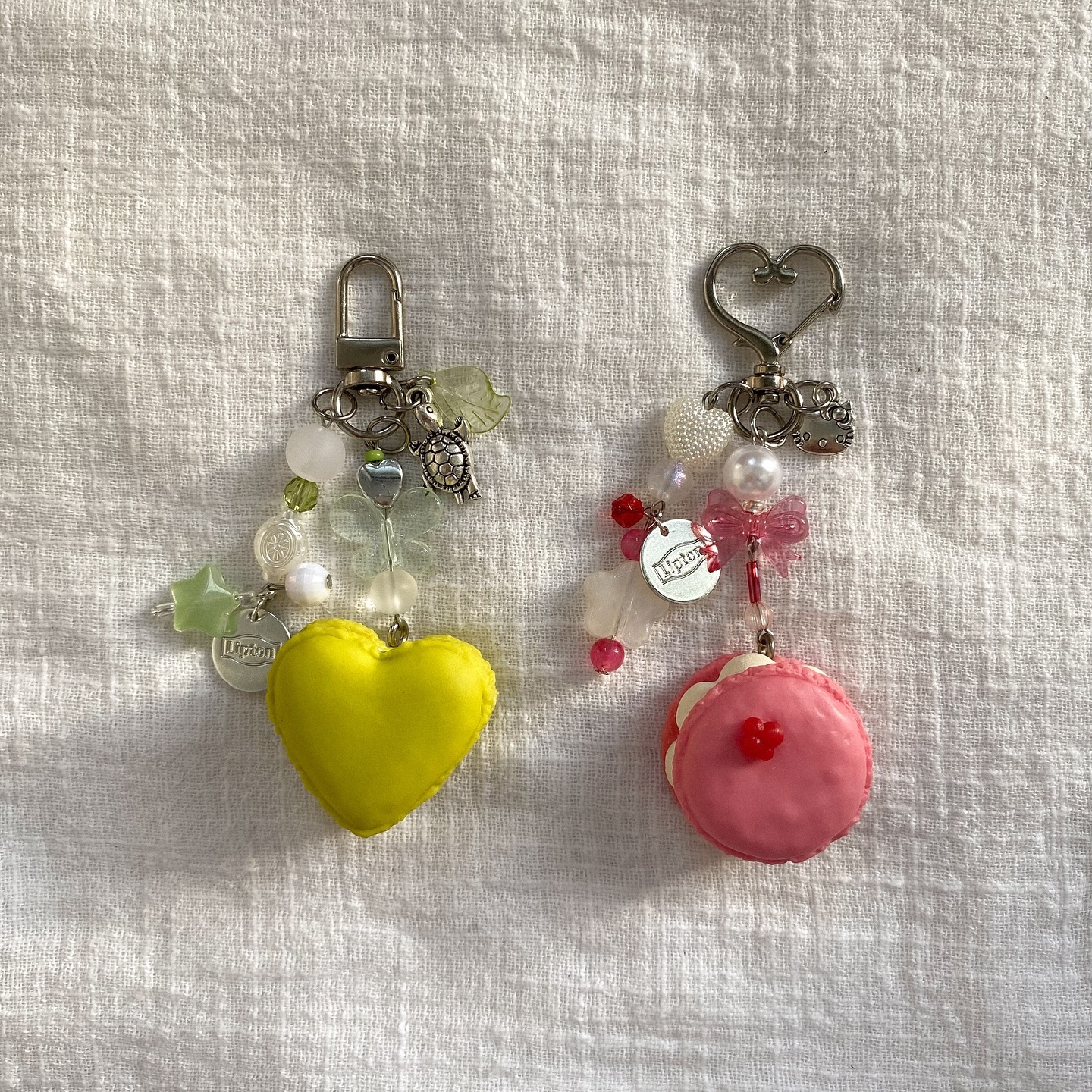 two keychains are shown on a white fabric background. the keychain on the left has a yellow heartshaped charm, a silver turtle charm, a silver lipton charm, and beads of various colors. the keychain on the right has a pink macaron charm, a pink bow charm, a silver lipton charm, and beads of various colors. both keychains have a silver clasp at the top.