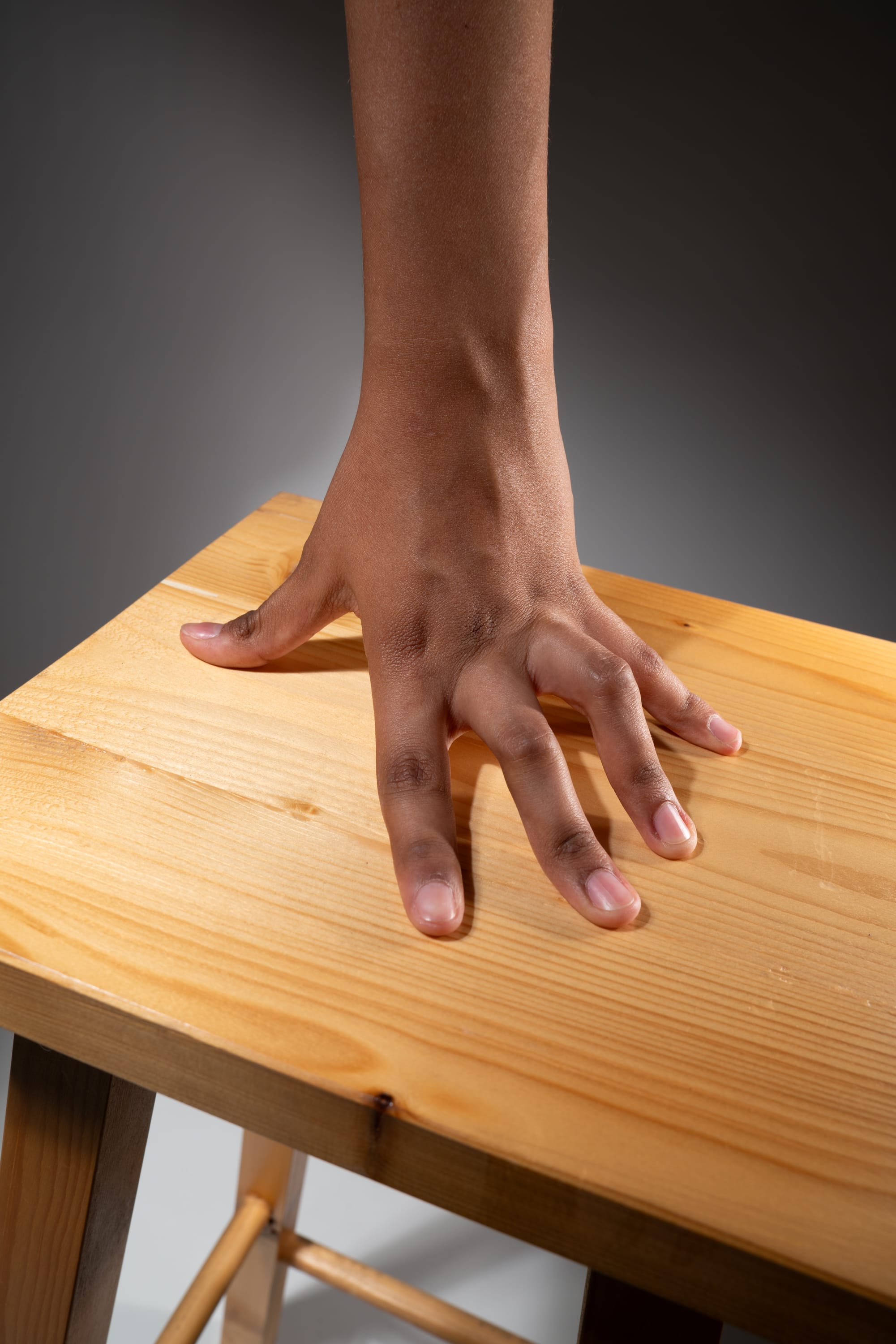 a closeup shot of a hand with brown skin pressing down on a light brown wooden table with its fingers spread out. the table is illuminated by studio lighting, creating a soft shadow under the hand. the perspective is from above, focusing on the hand and the texture of the wood.