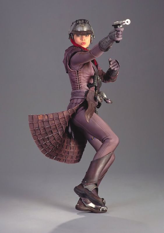 a female character from the star wars franchise, jyn erso from rogue one, is pictured in a fullbody shot standing with a blaster in hand.  jyn is a rebel fighter and is wearing a full costume and boots.  she is standing in front of a grey backdrop.