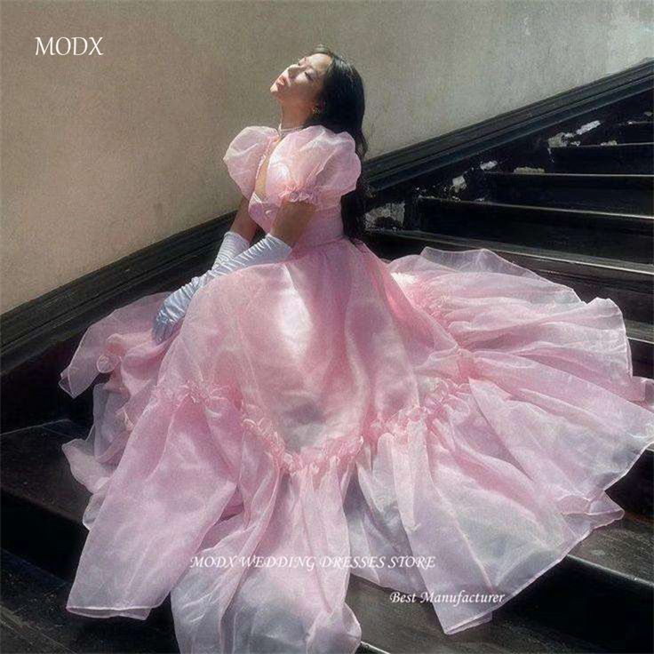 a woman in a long, flowing pink dress sits on stairs with her head tilted up. she is wearing long gloves and the dress has puffy sleeves. the dress appears to be made of organza or tulle. the words "modx" and "modx wedding dresses store best manufacturer" are visible in the image.