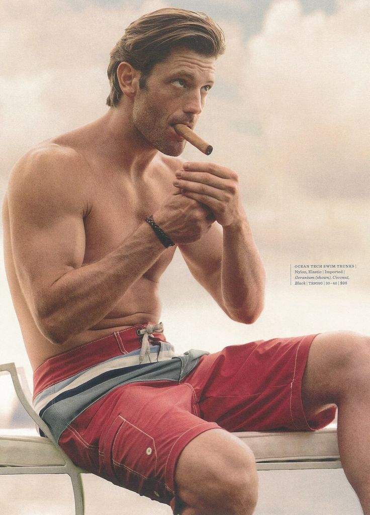 a man in swim trunks sits casually on a chair, holding a cigar in his hand. he is looking away from the camera. the man is shirtless, showing off his muscular physique. he is in a relaxed and casual pose, enjoying the summer weather. the man is wearing a pair of red and blue swim trunks. he is looking away from the camera. the image is a lifestyle shot that captures a moment of relaxation and enjoyment.