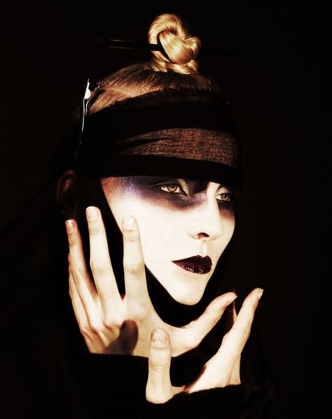 a woman with dark makeup and a mysterious expression stares intensely at the camera. her hair is styled in a bun and she is wearing a black band around her forehead.  she has dark eye makeup with a hint of blue and her lips are painted a dark purple. the image is in a dark setting.  the overall tone of the image is dramatic and mysterious.