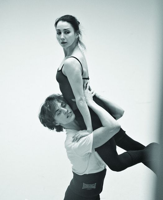 a black and white image of a male and female ballet dancer in a studio setting. the female dancer is being lifted by the male dancer, her legs are straight and she is looking over her shoulder with a serious expression on her face. the male dancer is looking at the ground with a focused expression on his face. both dancers are wearing black leotards and tights. this is a captivating image that captures the beauty and artistry of ballet.
