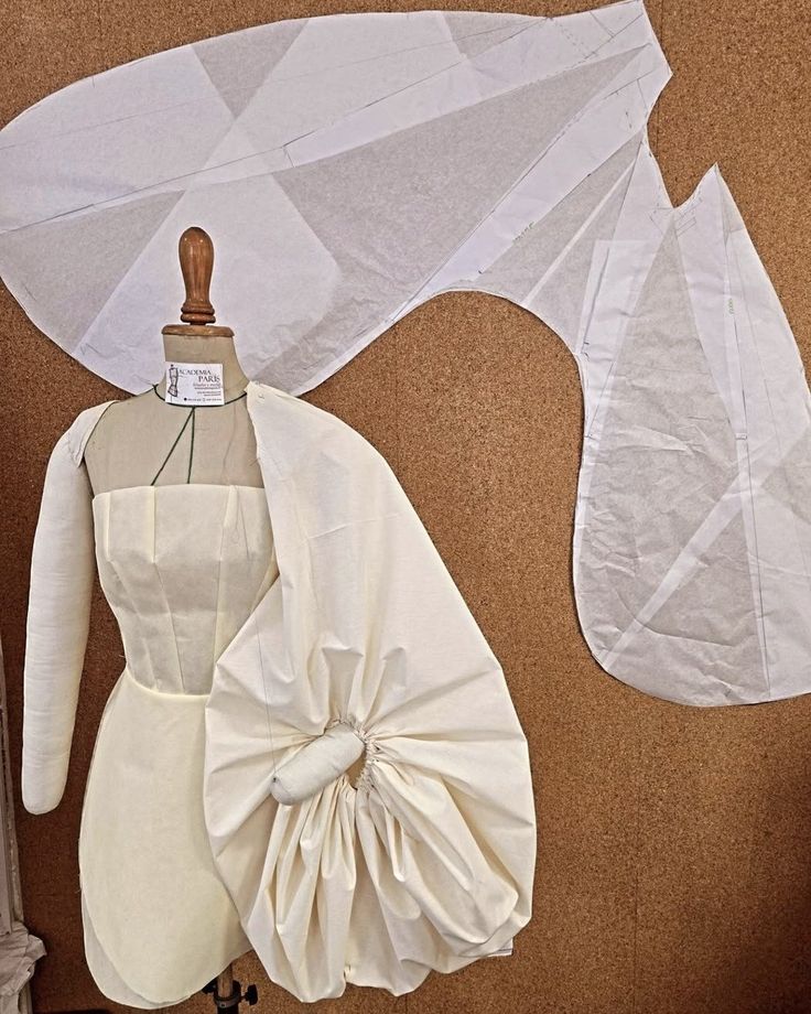 a dressmaker's dummy stands in front of a cork board. the dummy is wearing a white dress with one sleeve which is bunched up near the shoulder. above the dummy are two large paper sewing patterns for a sleeve.