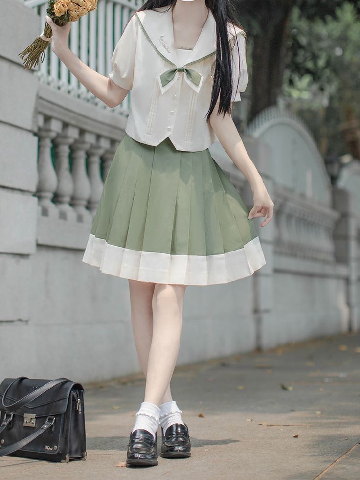 a young woman poses on a sidewalk while wearing a white and green school uniform style outfit. she is holding a bouquet of flowers in her right hand and a black purse hangs off her left shoulder. she is wearing white socks and black shoes. the background is out of focus and shows a building and some greenery.