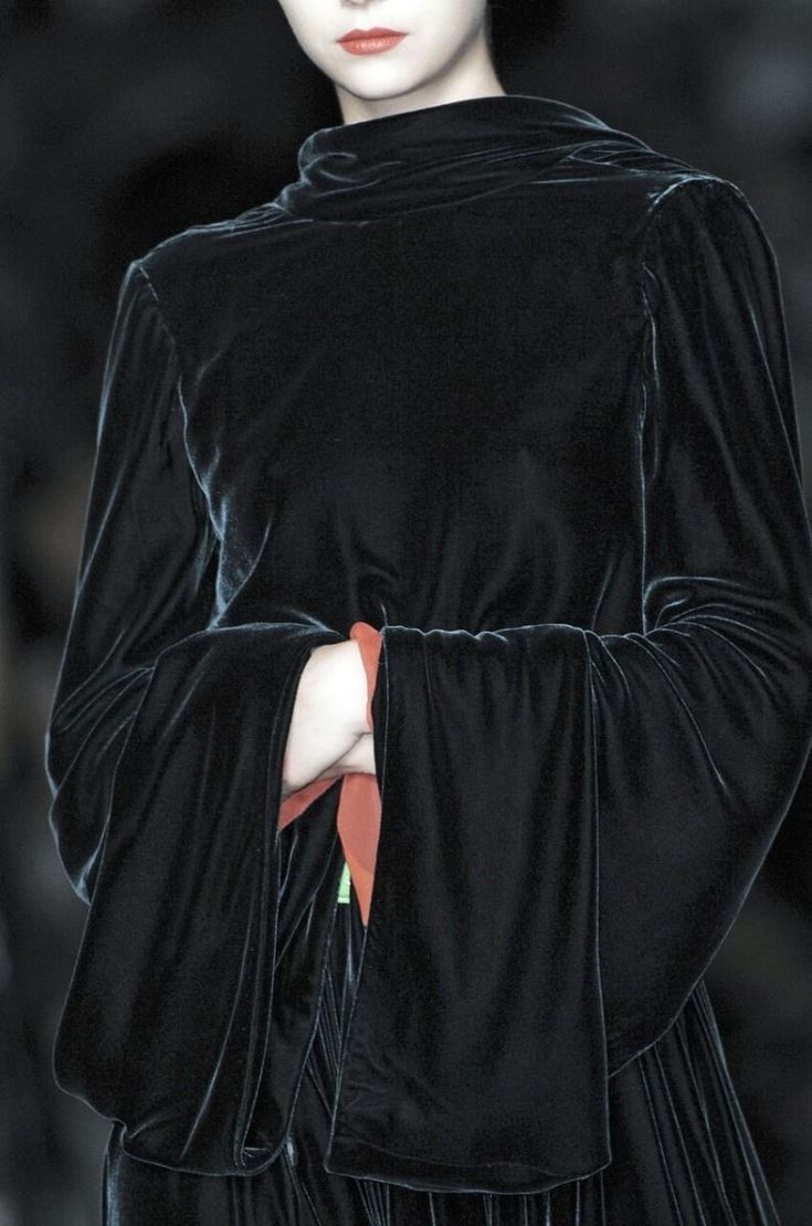 a woman in an allblack velvet dress is photographed in a studio. the sleeves are long and flowing, and the dress is belted at the waist. the woman looks directly at the camera with a serious expression.