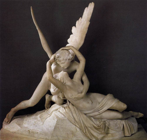 a white marble sculpture of cupid and psyche. cupid, the roman god of love, is depicted with delicate wings, kneeling and embracing psyche, a mortal woman who became his wife. psyche is partially draped in flowing fabric, her body relaxed in cupid's arms. they are positioned on a rocky base, suggesting a natural setting. the lighting is soft and highlights the smooth texture of the marble, emphasizing the tenderness of their embrace.