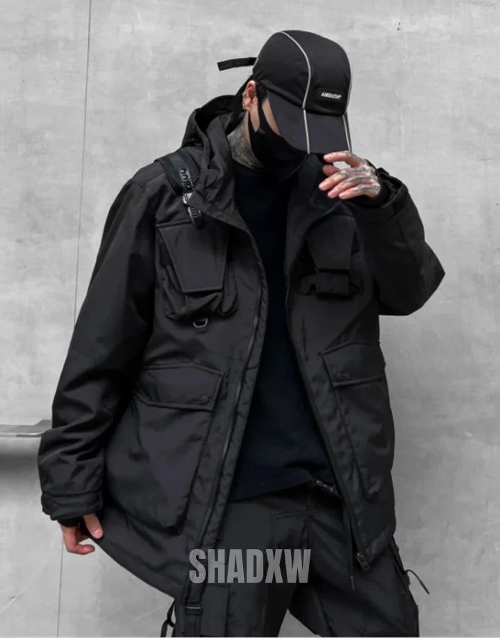 a young man is standing in front of a gray wall, posing for a photo. he is wearing a black jacket with multiple pockets, black pants and a black cap. he is looking down. the man is wearing a black mask and the word "shadxw" is written on the bottom of the image.