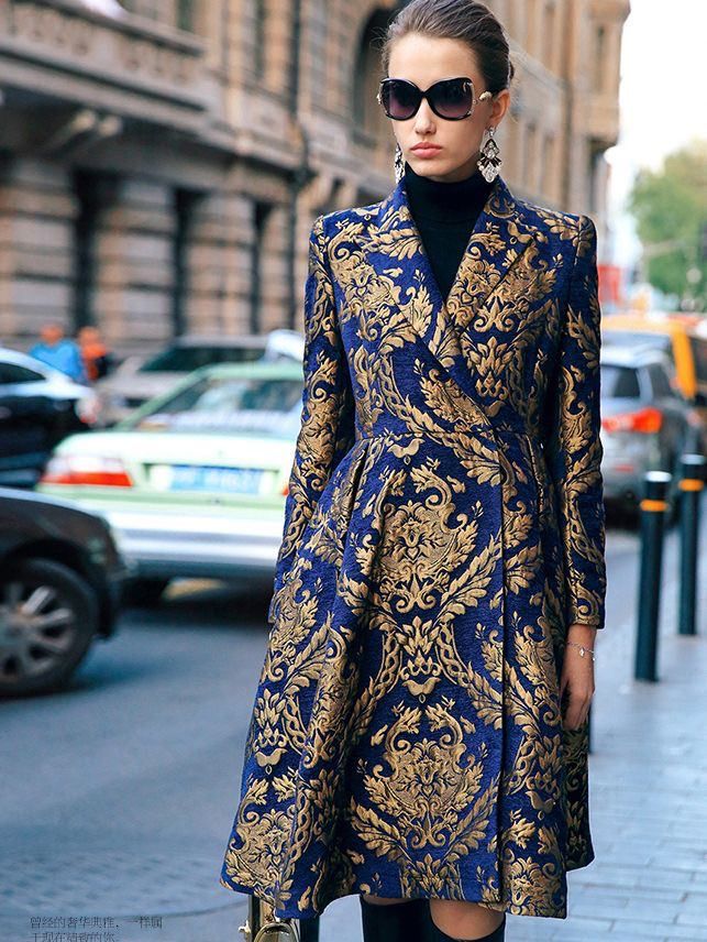 the image shows a stylish woman wearing a striking blue and gold brocade coat. the coat is a midilength, aline style with a doublebreasted closure. the fabric features a rich damask pattern in gold against a deep blue background. she's accessorized with oversized sunglasses, dangling earrings, and a black turtleneck. the setting appears to be a city street, with cars and buildings visible in the background. the overall aesthetic is one of elegant, highfashion street style.  the woman is posed midwalk, casually but stylishly presenting the outfit.