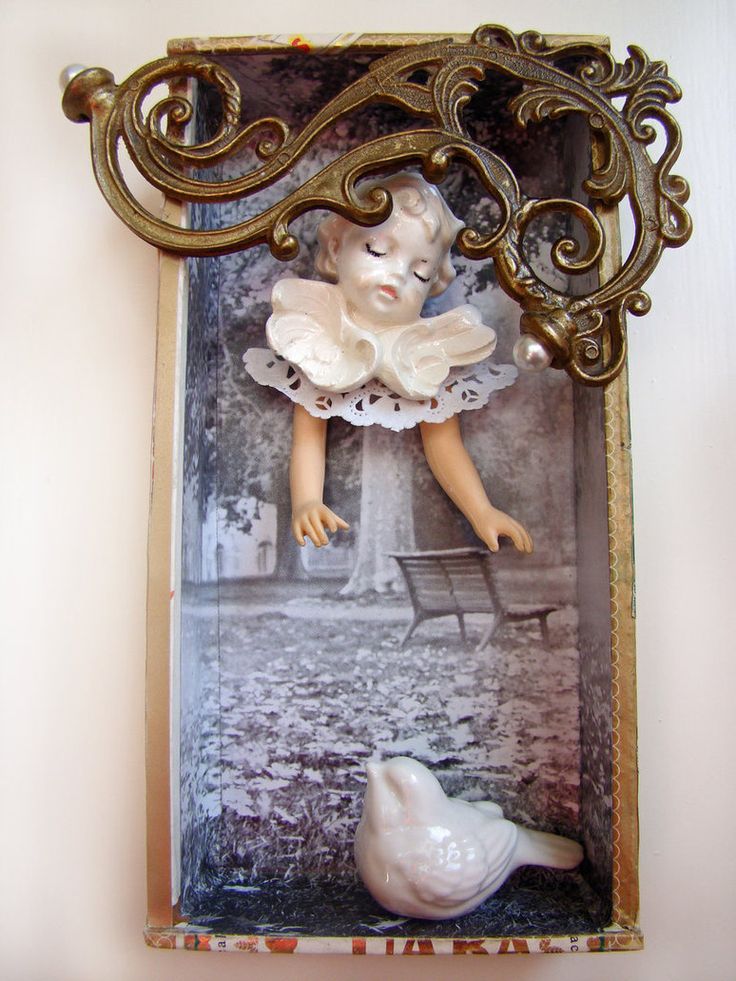 a shadow box featuring a porcelain doll figure suspended in front of a black and white park scene with a bench. a white ceramic bird is at the bottom of the box. an ornate metal ornament with pearl accents is affixed to the top of the box. the box itself is decorated with a patterned trim.