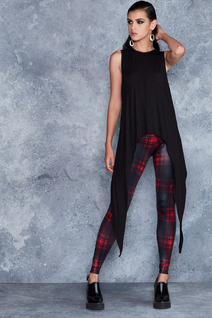 a woman is standing in front of a gray background wearing a black sleeveless top and plaid leggings. she is also wearing earrings and high heels.