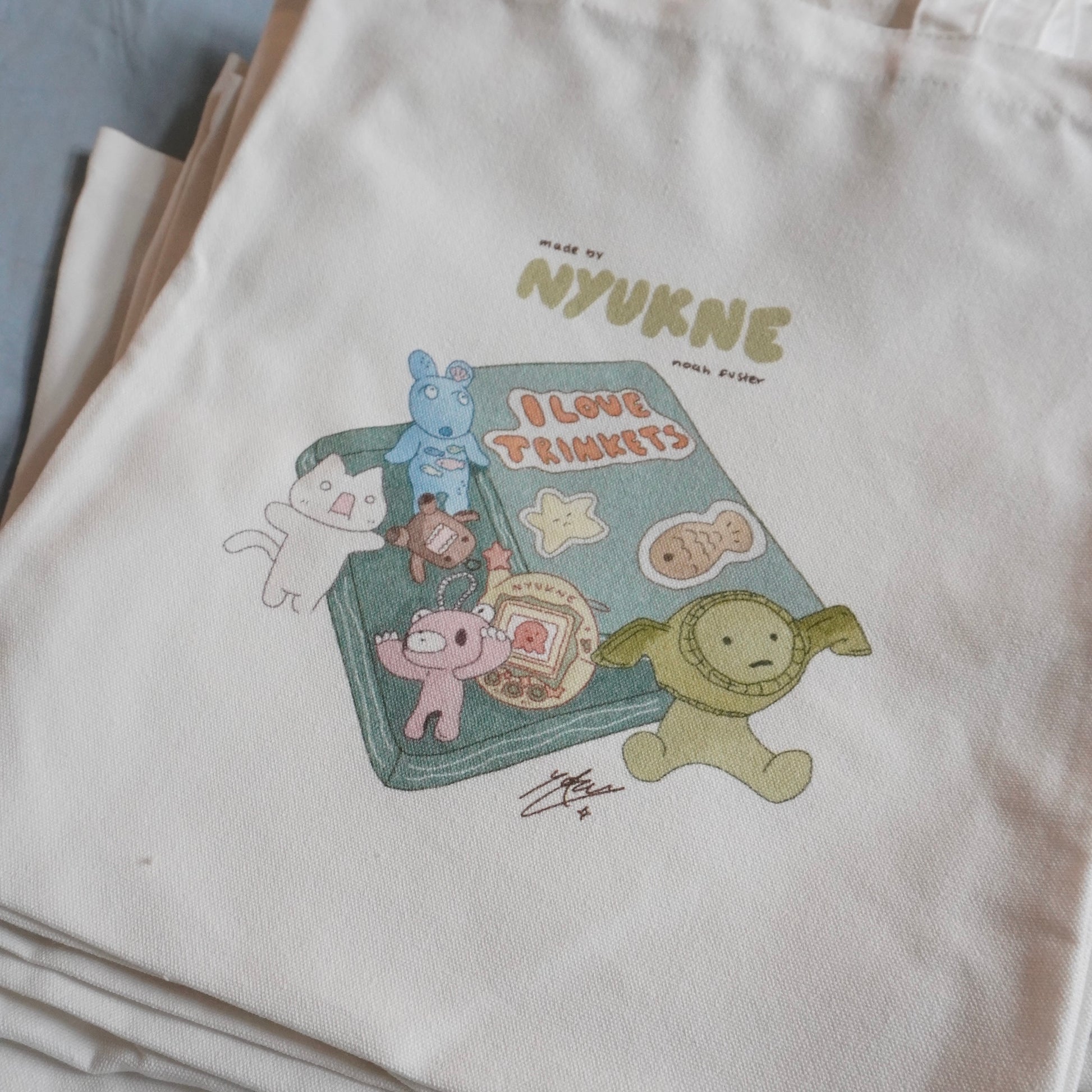 a closeup shot of a stack of beige canvas tote bags. the top bag features a charming illustration of a book titled "i love trinkets," surrounded by various cute characters like a bunny, cat, fish, and monsters in pastel colors. the bag is labeled "made by nyukne noah fisher," indicating the creator's name. the overall style of the illustration is cute, kawaii, and reminiscent of children's book illustrations. the bags are stacked neatly, showcasing the print clearly on the topmost bag.