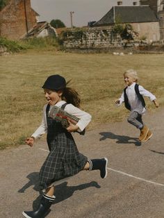 two young children, a girl and a boy, are captured midrun on a paved road. they are both dressed in school uniforms, with the girl sporting a black and green plaid dress and the boy wearing a white shirt with a black vest and grey pants. the girl carries a book and they both wear backpacks. their joyous expressions and dynamic poses convey a sense of freedom and playfulness. the background features a picturesque rural setting, with fields, trees, and old buildings. the overall tone of the image is warm and nostalgic, evoking a sense of carefree childhood memories.