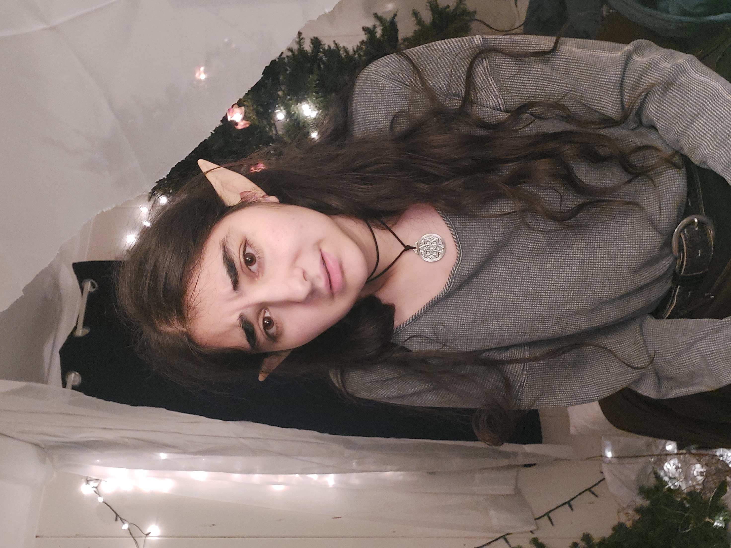 a closeup portrait of a young woman with long, dark, wavy hair.  she's wearing elf ears, a grey waffleknit sweater, a dark necklace with a circular pendant, and a black belt.  she has a slightly concerned or pensive expression on her face.  the background includes blurry christmas lights and a partially visible christmas tree, suggesting a holiday or winter setting.  the overall aesthetic leans towards fantasy or cosplay.